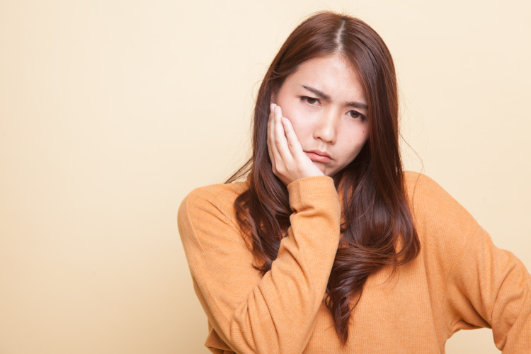 Brunette woman in a yellow sweater cringes in pain and cradles her jaw due to TMJ dysfunction