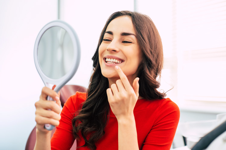 Brunette woman smiles in a handheld mirror after transforming her smile with cosmetic dentistry