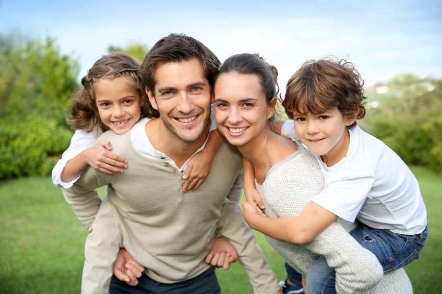 A smiling young family with a dad and mom each carrying a young child piggyback.