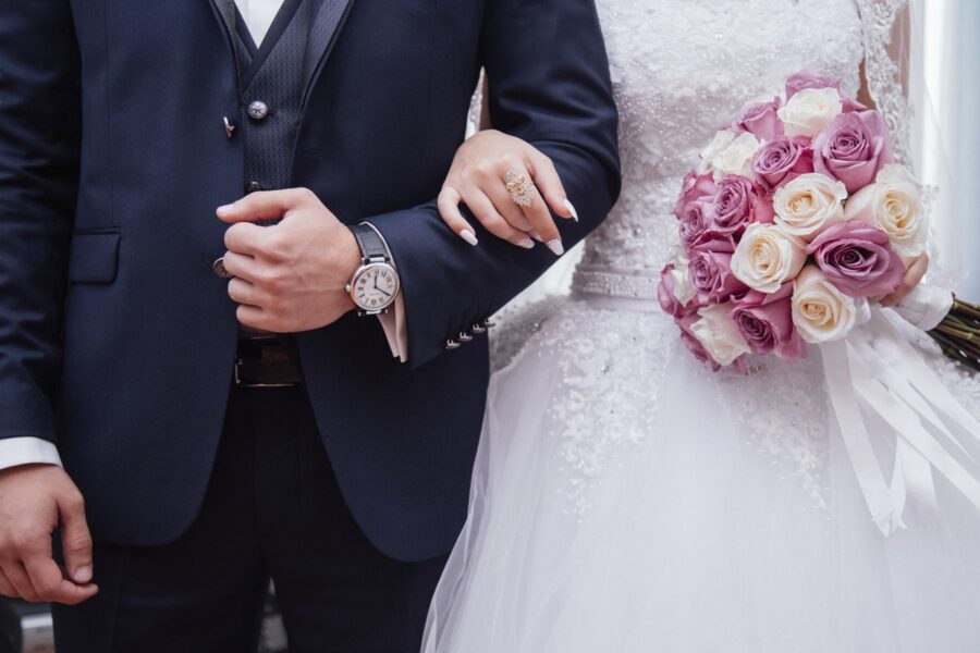 Chests down image of bride holding a pink rose bouquet and the arm of her groom in a handsome suit