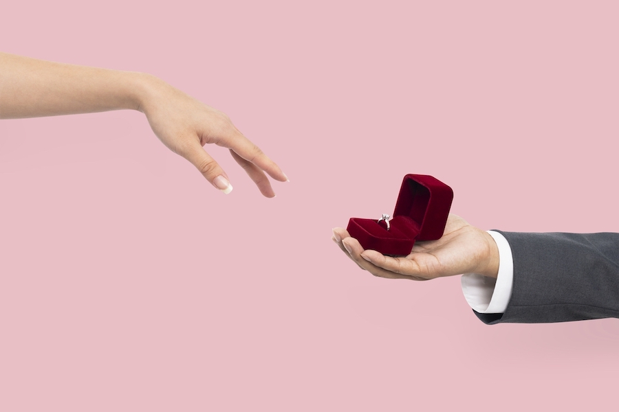 A woman reaches her manicured hand toward a man holding an engagement ring in a velvet box