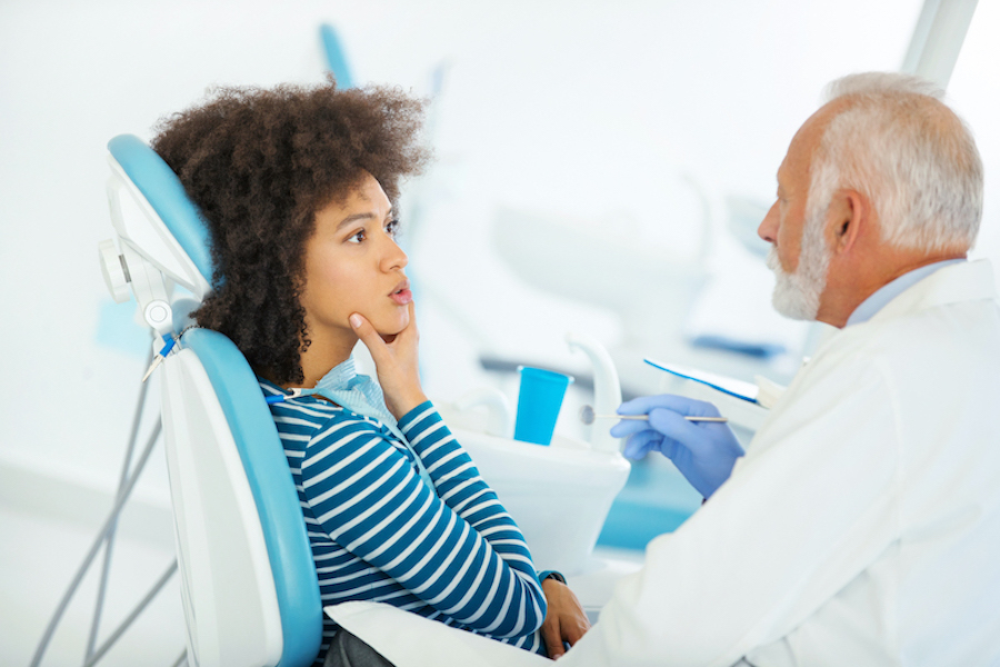 Black woman touches her jaw as she sits in the dental chair talking with her dentist about tooth replacement