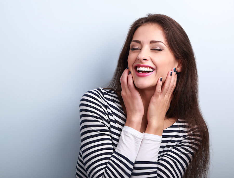 Brunette woman in a black and white striped shirt smiles and touches her cheeks after receiving rejuvenating dermal fillers